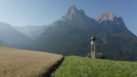 Secluded-Nature-grass-and-wheat-field-with-a-small-church-in-Europe-Mountain-Backdrop-French-Alps,-Kastelruth,-Trentino