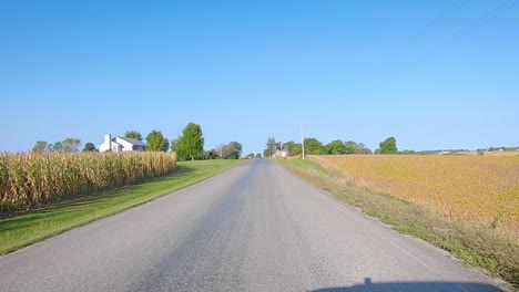 POV-driving-on-rural-county-road-past-maturing-fields,-farmyard,-and-bicyclist-in-rural-Iowa-on-a-sunny-early-autumn-day