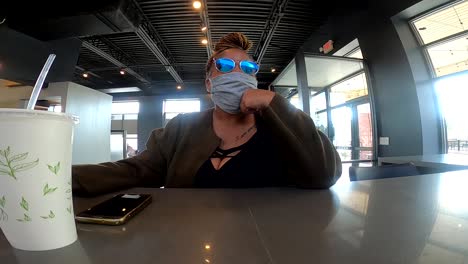 Schwarze-Frau-Mit-Maske-Sitzt-Im-Restaurant-Und-Wartet-Darauf,-Mit-Schützender-Gesichtsmaske-Und-Sonnenbrille-Zu-Bestellen