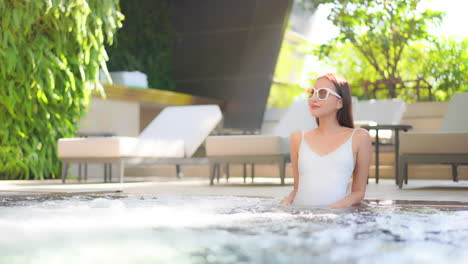 Fancy-Asian-Female-in-Sunglasses-and-Swimsuit-Enjoying-in-Jacuzzi-of-a-Swimming-Pool-on-Sunny-Summer-Day,-Slow-Motion