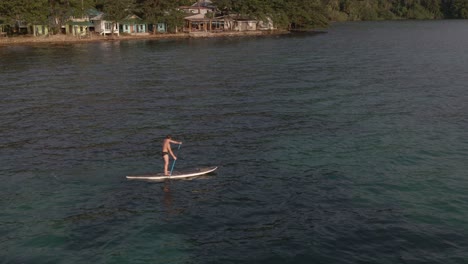Aerial-drone-bird's-eye-view-of-man-exercising-on-a-sup-paddle-board-in-turquoise-tropical-clear-waters,-with-rocky-coastline-and-backpacker-style-bungalows-in-Koh-Chang-Thailand