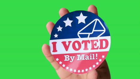 Hand-of-a-man-holding-I-Voted-By-Mail-sticker-against-green-screen-chroma-key