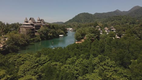 Drone-birds-eye-view-of-a-tropical-river-with-a-big-luxury-resort-and-mountains-with-paddle-boarders-paddling-down-the-river-on-the-Island-of-Koh-Chang,-Thailand