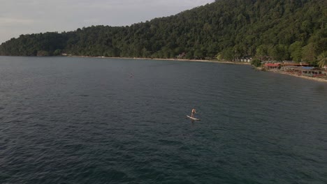 Aerial-drone-bird's-eye-view-quick-moving-backwards-ascending-dolly-shot-of-man-exercising-on-a-sup-paddle-board-in-turquoise-tropical-clear-waters,-with-beach-and-coastline-in-Thailand