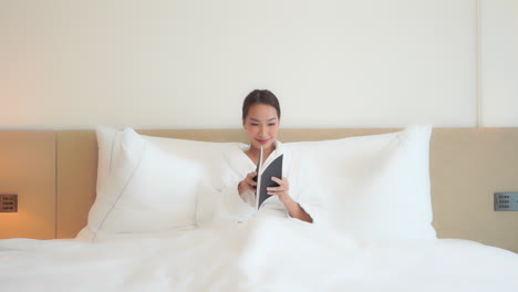 Asian-Beauty-Reading-a-Book-in-a-Hotel-Bed,-Clean-White-Room-SLOMO