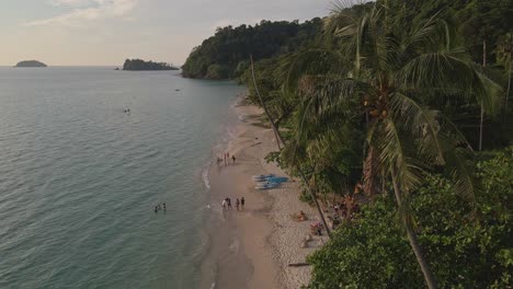 Tropical-Island-drone,-bird’s-eye-view-static-shot-with-lush-green-rain-forest-and-tropical-palm-trees-with-white-sand-beach-and-small-beach-bars-and-restaurants-and-bungalows-with-domestic-tourists