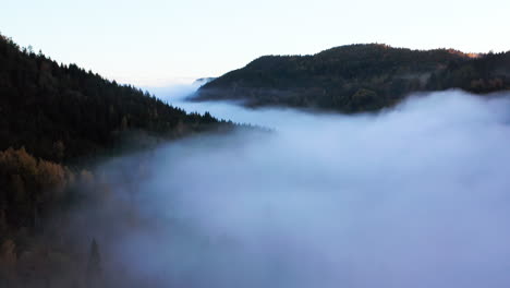 Thick-fog-hovers-over-forest-mountains-in-Norway,-Aerial-Drone-Shot