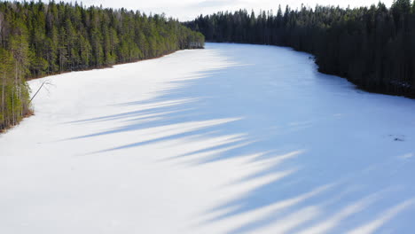 Drone-shot-of-remote-narrow-icy-lake-and-nice-shadows-from-spruce-trees-on-the-ice