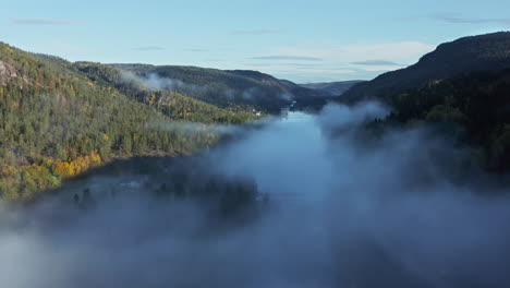 Aerial-drone-view-over-a-lake-and-waterfall,-in-middle-of-hills-and-foliage-forest,-foggy,-autumn-morning,-in-South-Norway
