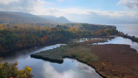 flying-over-marshlands,-a-river,-and-fall-foliage-toward-distant-mountains-and-hills