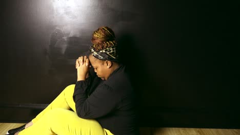 Sideview-of-Black-woman-sitting-on-floor-against-black-wall-praying-to-God