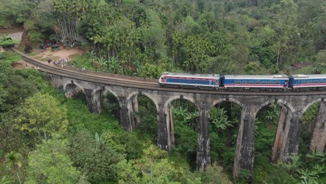 blue-train-crossing-the-Nine-Arches-Bridge-in-Sri-Lanka-surrounded-by-lush-green-landscape,-tea-plantations-and-rolling-hills-Cinematic-Drone-Aerial-in-4K