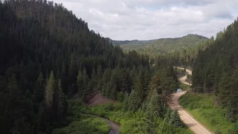 Aerial-shot-moving-through-a-mountain-forest-valley-with-a-dirt-road-and-a-stream,-revealing-a-wooden-trestle-bridge-on-the-Mickelson-Trail-in-the-Black-Hills,-South-Dakota