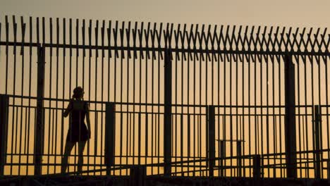 Silhouette-of-Young-Woman-Walking-in-Front-of-Fence-during-Sunset