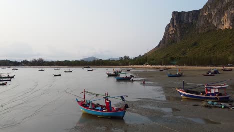 AERIAL:-Asian-Fishing-Boats-stranded-at-low-tide-on-the-coast-with-rocky-mountains-on-the-background-in-Thailand