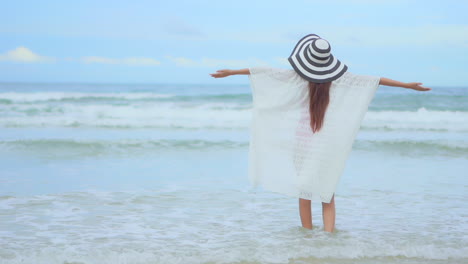 Asiatische-Frau,-Die-Im-Sommer-Am-Strand-Am-Wasser-Steht-Und-Ein-Sommerkleid-Und-Einen-Gestreiften-Hut-Trägt