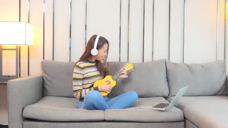 A-pretty-young-woman,-wearing-headphones,-sits-on-a-comfortable-sofa-learning-to-play-the-ukulele-from-an-online-source