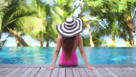 Vista-Trasera-De-La-Mujer-Sentada-Al-Borde-De-La-Piscina-En-Un-Hotel-Exótico-En-Bahamas-Con-Monokini-Rosa-Y-Sombrero-A-Rayas