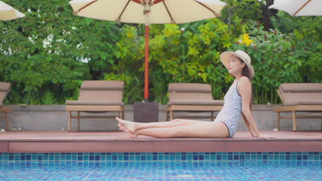Asian-model-woman-sitting-stretching-her-legs-at-the-edge-of-the-swimming-pool-daytime-in-Thailand-resort,-side-view
