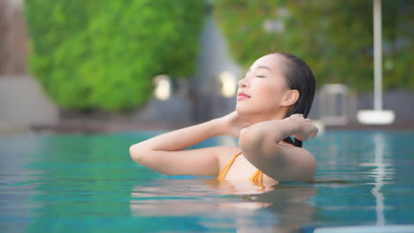 Portrait-of-Beautiful-brunette-asian-woman-bathing-in-a-swimming-pool,-she-is-in-water-up-to-the-neck-and-touching-her-hair-face-close-up-slow-motion-side-view