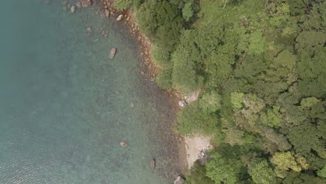Aerial,-birds-eye-view-of-tropical-rocky-granite-ocean-coastline-,-clear-turquoise-water-with-small-beach,-lush-tropical-forest-vegetation-on-a-Island