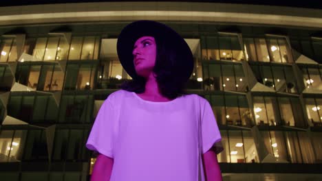 Woman-In-Hat-Under-Purple-Light-Looking-From-Side-To-Side-With-Futuristic-Building-In-Background---Low-Angle-Shot