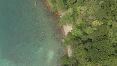 Aerial,-birds-eye-view-of-tropical-rocky-granite-ocean-coastline-with-clear-blue-ocean-and-small-beach-with-lush-tropical-forest-vegetation-on-a-Island