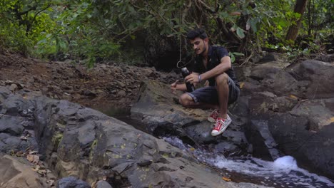 Junge-Indische-Füllwasser-Aus-Natürlichem-Fluss-Trockenfluss-Im-Dschungel-Indien-Trägt-Shorts