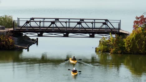 An-aerial-view-of-two-people-slowly-kayaking-away-from-the-camera-and-going-under-a-pedestrian-bridge