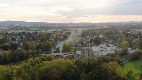 Manufacturing-mill-with-bins-and-towers,-smoke,-steam-rising-from-exhaust-pipes-in-small-town-in-USA