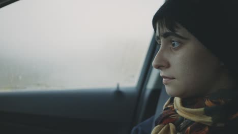A-Sad-Face-Woman-In-Brown-Scarf-And-Black-Beanie-Looking-Outside-A-Foggy-Car-Window---Closeup-Shot-Handheld