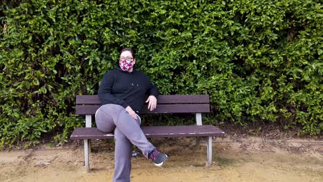 Woman-with-face-mask-sits-on-park-bench-looking-around-and-at-camera-in-pandemic