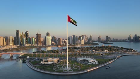 Luftaufnahme-Der-Flagge-Der-Vereinigten-Arabischen-Emirate,-Die-In-Der-Luft-Weht,-Der-Blaue-Himmel-Im-Hintergrund,-Das-Nationale-Symbol-Der-Vae-über-Der-Flaggeninsel-Von-Sharjah,-Vereinigte-Arabische-Emirate,-4k-video
