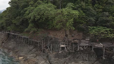 Drone-left-trucking-orbit-shot-,-birds-eye-view-of-a-broken-down-wooden-walkway-along-a-granite-rock-coastline-on-a-tropical-Island-with-jungle-lush-green-forest-and-ocean