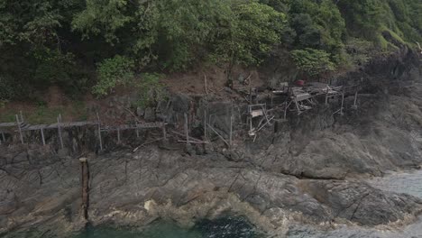 Drone-trucking-orbit-shot-,-birds-eye-view-of-a-abandoned-derelict-wooden-board-walk-and-deck-along-granite-rock-coastline-on-a-tropical-Island-with-jungle-lush-green-forest-and-ocean