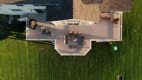 Rising-aerial-top-down-view-of-large-deck,-outdoor-living-space-with-white-railing,-table,-chairs,-grill-among-green-grass-yard