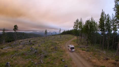 Drone-follows-a-camper-van-down-a-dirt-road-in-the-mountain-at-sunset-during-wildfires