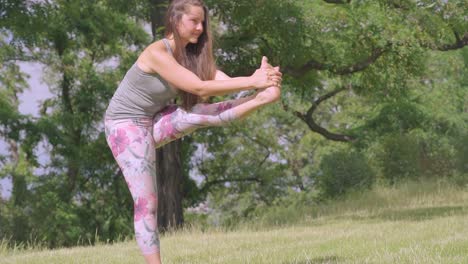 Caucasian-woman-holds-leg-yoga-pose-in-nature,-slow-motion-tilt-shot