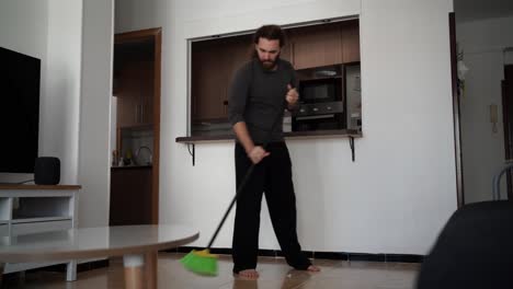 Video-of-a-bearded-man-sweeping-his-living-room's-floor-with-a-black-and-green-broom