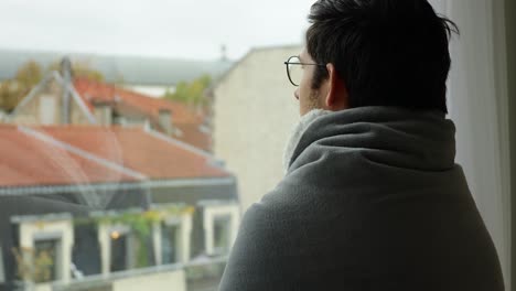 Young-man-wearing-hot-blanket-and-looking-through-a-window-during-overcast-day