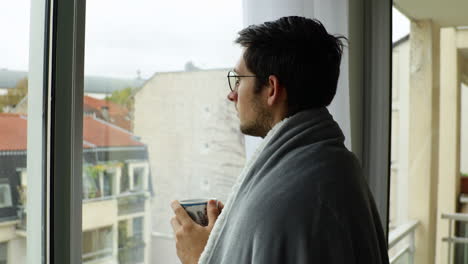 Young-man-staring-at-window-and-drinking-coffee-while-wearing-cosy-blanket-during-overcast-day
