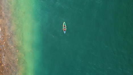 Birdseye-Overhead-shot-of-Two-People-in-Kayak-Paddling-across-Turquoise-Green-Exotic-Shallow-Ocean-Waters,-Drone-Aerial