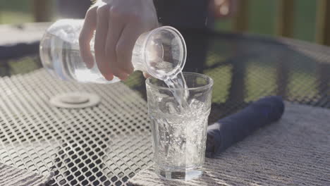 Hand-pouring-water-with-ice-cubes-from-jug-into-glass-on-outdoor-table