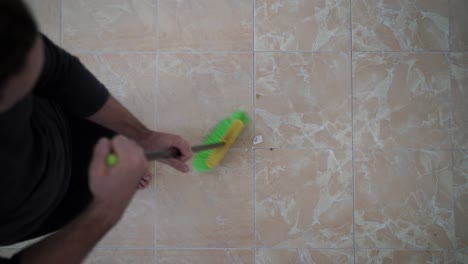 Video-from-above-of-a-bearded-man-sweeping-his-living-room's-floor-with-a-black-and-green-broom