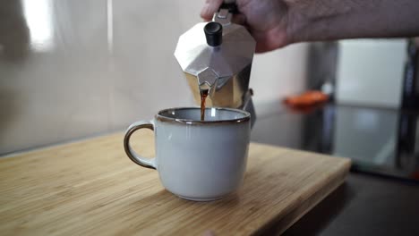 Video-Einer-Hand,-Die-Kaffee-Aus-Einer-Italienischen-Kaffeemaschine-Direkt-In-Eine-Kaffeetasse-Auf-Einem-Holzschneidebrett-In-Der-Küche-Gießt