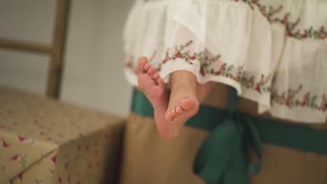 Cute-Girl-dressed-in-festive-dress-sitting-on-Christmas-Gifts-swinging-her-feet---close-up-shot