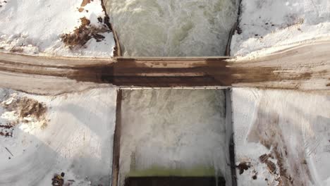 Bird's-eye-view-of-small-Dam-icy-floodgate,-flowing-through-cold-white-stream---Aerial-shot