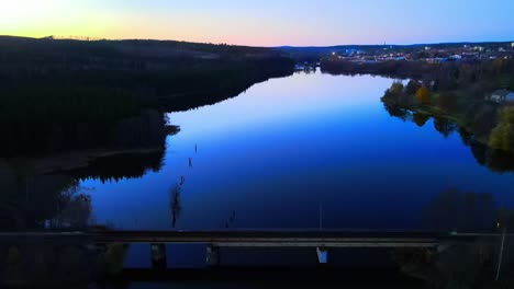 Drone-footage-revealing-a-train-bridge-on-a-beautiful-evening-with-calm-lake-in-the-foreground-and-the-sunset-in-the-background