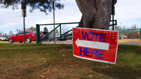 Vote-Here-Sign-Pointing-Left-with-People-Driving-Cars-in-Parking-Lot-Background