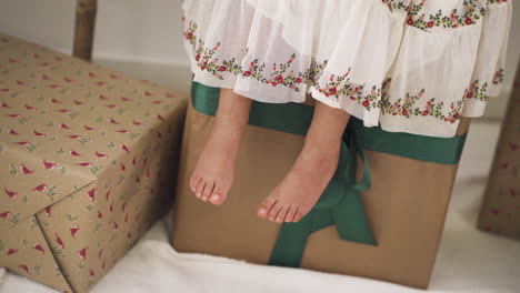 Girl-swinging-her-feet-dressed-in-festive-dress-sitting-on-Big-Christmas-Gift-Box---High-angle-close-up